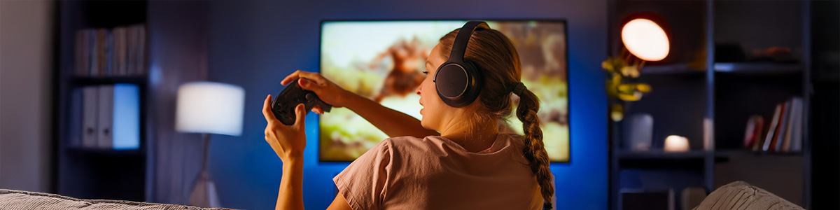 Osoba w słuchawkach gra na konsoli przed telewizorem. W pokoju regały i lampa.
