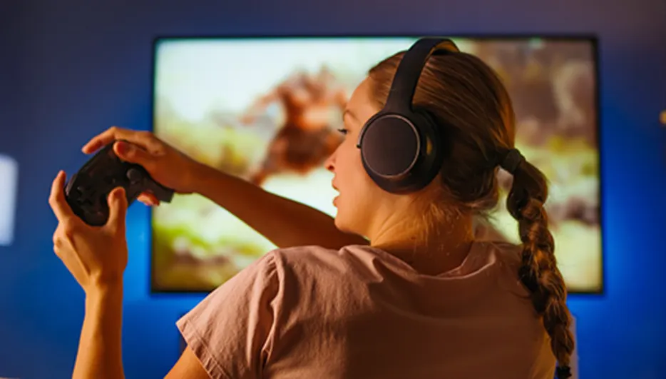 Osoba w słuchawkach gra na konsoli przed telewizorem. W pokoju regały i lampa.