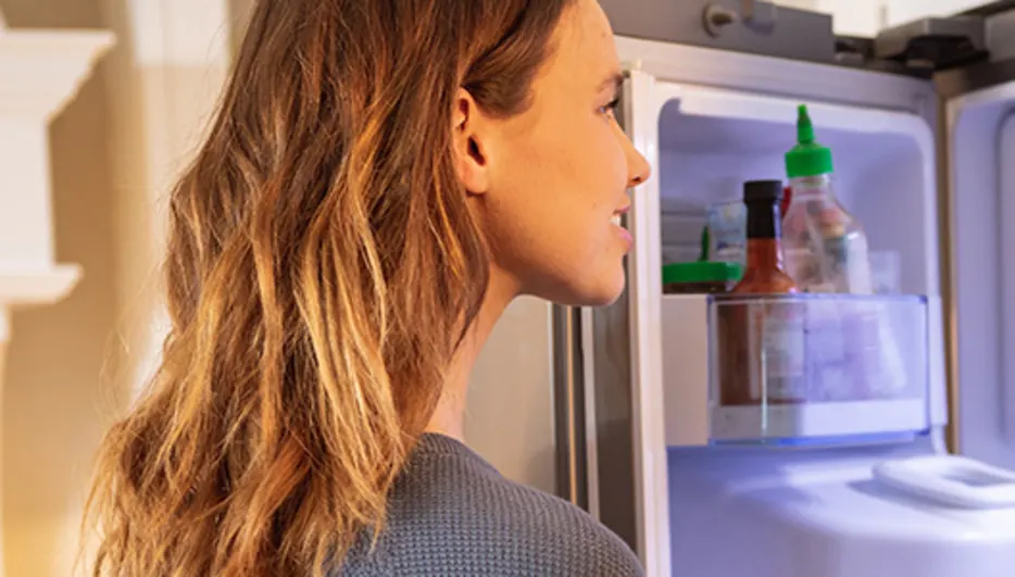 Kobieta stoi przed otwartą lodówką. Wewnątrz widać butelki z sosem. W tle białe szafki kuchenne.