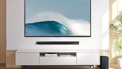 Telewizor na ścianie nad białą szafką RTV. Pod telewizorem czarny soundbar. Z boku stoi doniczka z zieloną rośliną.