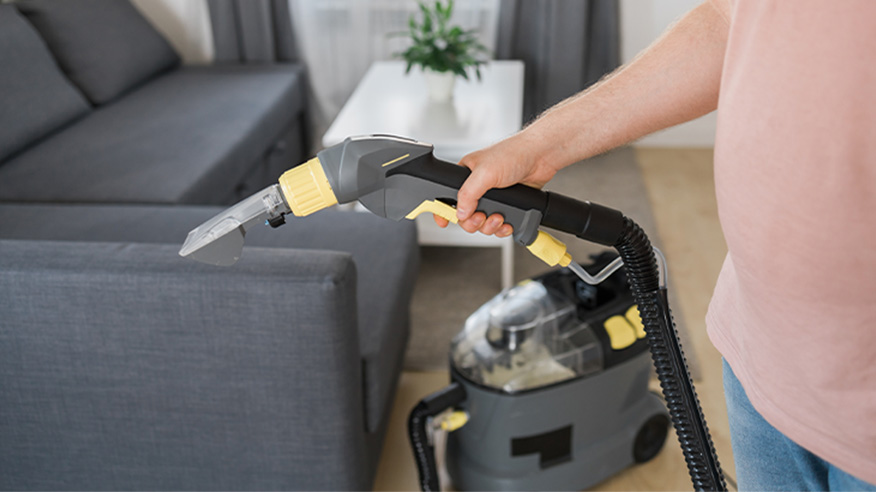 Odkurzacz piorący z małą ssawką podczas pracy przy kanapie z ciemną tapicerką.