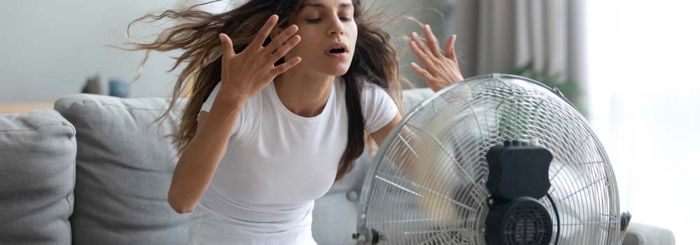 Kobieta w białej koszulce stoi przed wiatrakiem w pokoju. Za nią szara sofa i okno.