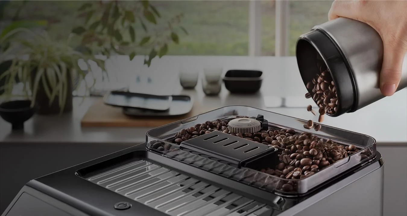 Osoba wsypuje ziarna kawy z metalowego pojemnika do przezroczystego zbiornika w ekspresie do kawy. W tle widać rośliny i naczynia.