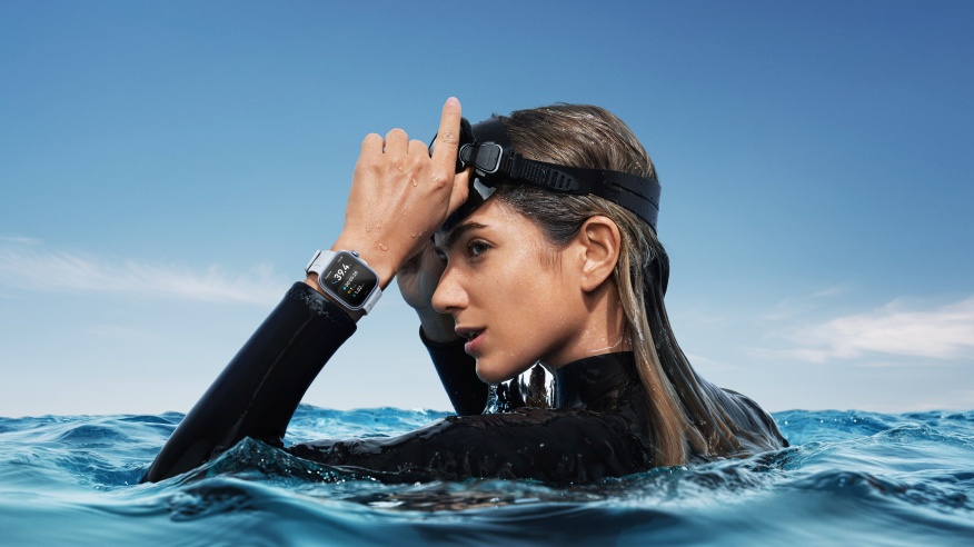 Kobieta w wodzie w czarnym stroju pływackim i goglach. Na nadgarstku zegarek. W tle błękitne niebo i woda z falami.