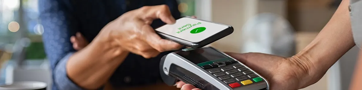 Mężczyzna w koszuli płaci zbliżeniowo telefonem przykładając smartfon do terminala płatniczego.