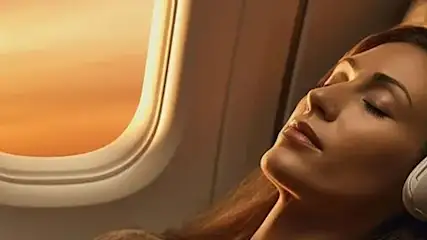 Na zdjęciu widać śpiącą kobietę w słuchawkach w fotelu samolotu, obok okna z pomarańczowym światłem.