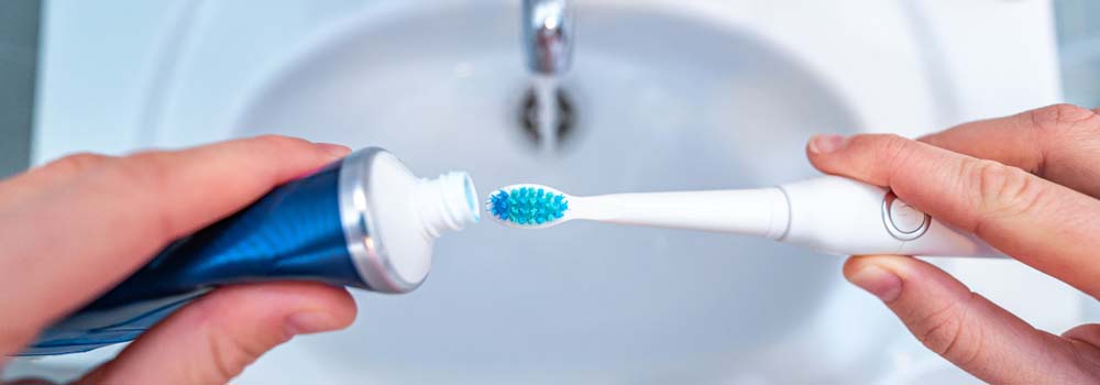 Osoba wyciska pastę z tubki na niebieską końcówkę szczoteczki elektrycznej nad białą umywalką z kranem.