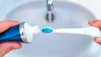 Osoba wyciska pastę z tubki na niebieską końcówkę szczoteczki elektrycznej nad białą umywalką z kranem.