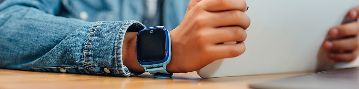 Dziecko w dżinsowej koszuli używa tabletu, na nadgarstku ma niebieski smartwatch, siedząc przy drewnianym stole.