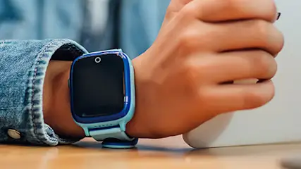 Dziecko w dżinsowej koszuli używa tabletu, na nadgarstku ma niebieski smartwatch, siedząc przy drewnianym stole.