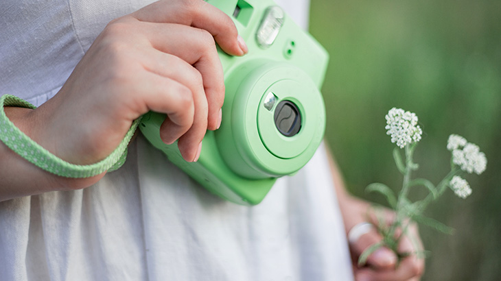 Osoba trzyma zielony aparat i białe kwiaty na tle zieleni. Aparat ma pasek. Osoba jest ubrana w białą koszulę.