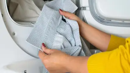 Osoba w żółtej bluzce wyjmuje z białej suszarki do ubrań szarą koszulę w kratkę.