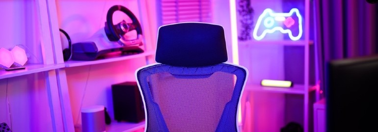 Krzesło biurowe stoi przodem do regału z neonem kontrolera gier, słuchawkami i kierownicą. Kolorowe oświetlenie.