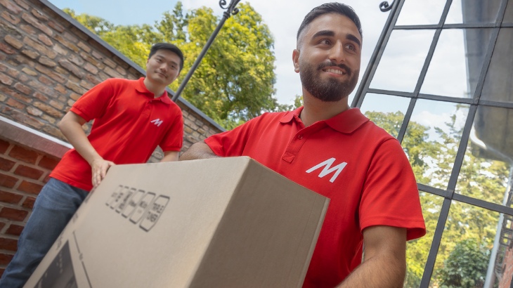 Dwóch mężczyzn w czerwonych koszulkach z logo niesie karton przed budynkiem z cegły i oknami.