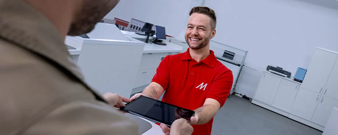 Uśmiechnięty mężczyzna w czerwonej koszulce z logo wręcza tablet klientowi. W tle białe meble biurowe i sprzęt.