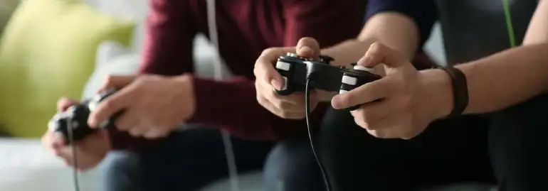 Dwie osoby grają w grę wideo, trzymając czarne kontrolery. W tle widać kanapę i poduszkę.