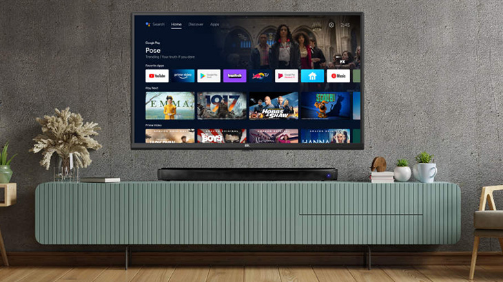 Telewizor na ścianie nad szafką RTV, na której stoi głośnik, obok rośliny doniczkowe i krzesło.