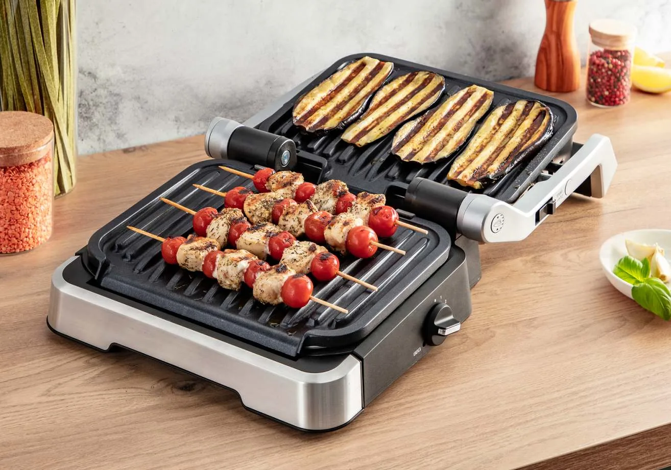 Na stole stoi elektryczny grill z pieczonymi szaszłykami z kurczaka z pomidorami i grillowanym bakłażanem.
