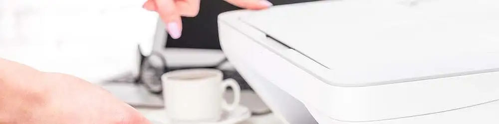 Osoba obsługuje białe biurowe urządzenie wielofunkcyjne. W tle widoczne filiżanka z kawą i laptop.