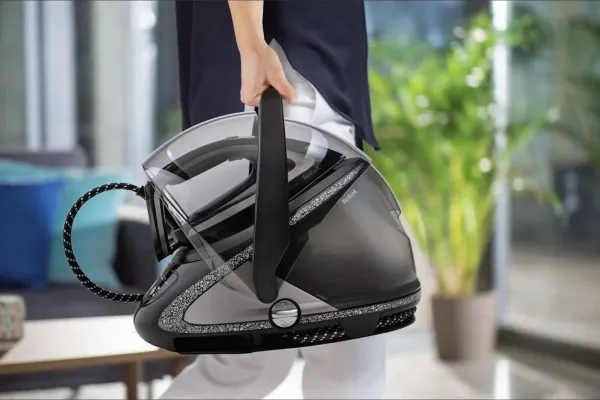 Osoba trzyma w ręku generator pary do żelazka, czarno-srebrny, z kablem w oplocie. W tle okno i roślina doniczkowa.