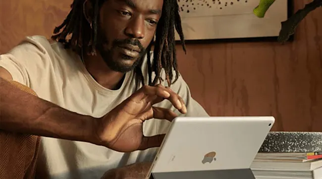 Mężczyzna używa tabletu. Za nim drewniana ściana i grafika. Tablet stoi na książkach obok rośliny.