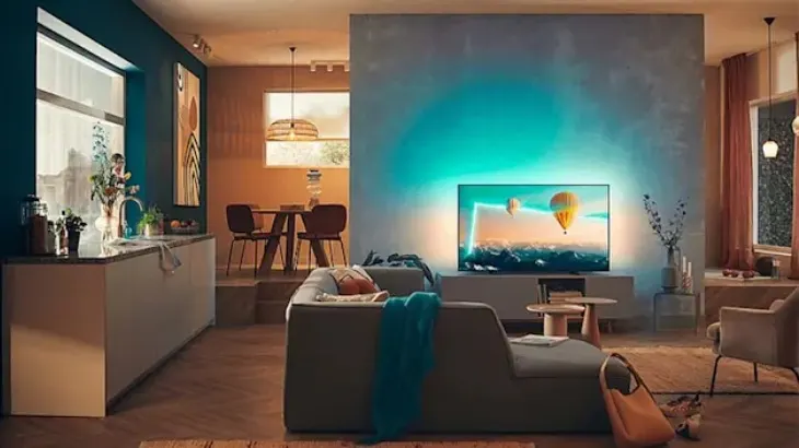 Salon z szarą sofą, telewizorem i drewnianym stołem. W tle jadalnia i kuchnia. Ściany w odcieniach błękitu i beżu, drewniana podłoga.