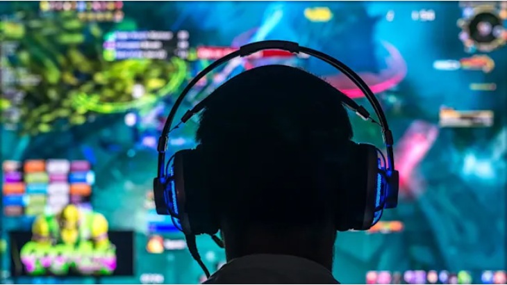 Osoba w słuchawkach przed monitorem wyświetlającym dynamiczną grafikę gry z elementami zielonymi, różowymi i niebieskimi.