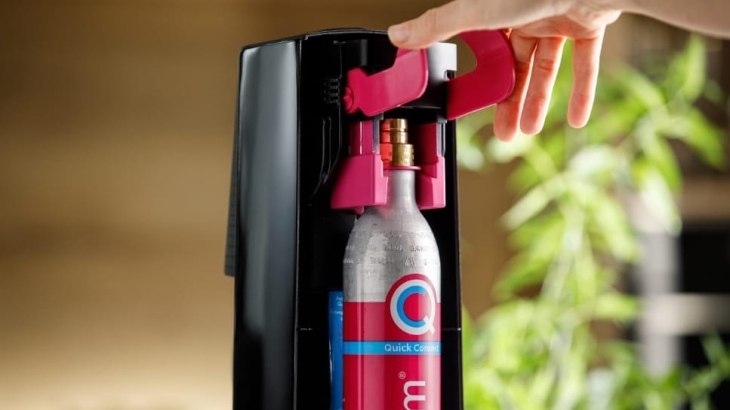 Dłoń obsługuje czarny saturator z czerwoną dźwignią i srebrno-czerwoną butlą CO2. W tle brązowa ściana i zielona roślina.
