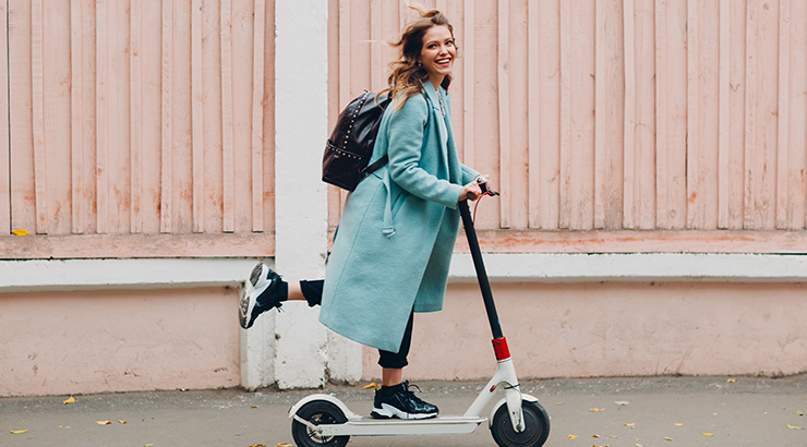 Kobieta w płaszczu jedzie na hulajnodze elektrycznej obok drewnianego płotu. Ma na sobie plecak.