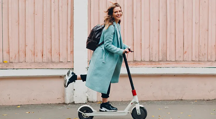Kobieta w płaszczu jedzie na hulajnodze elektrycznej obok drewnianego płotu. Ma na sobie plecak.