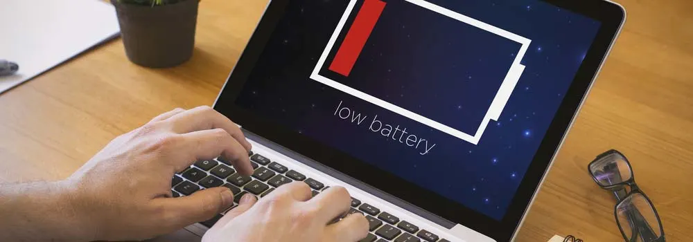 Laptop wyświetla komunikat o niskim poziomie baterii. Dłonie piszą na klawiaturze. Obok leżą okulary.