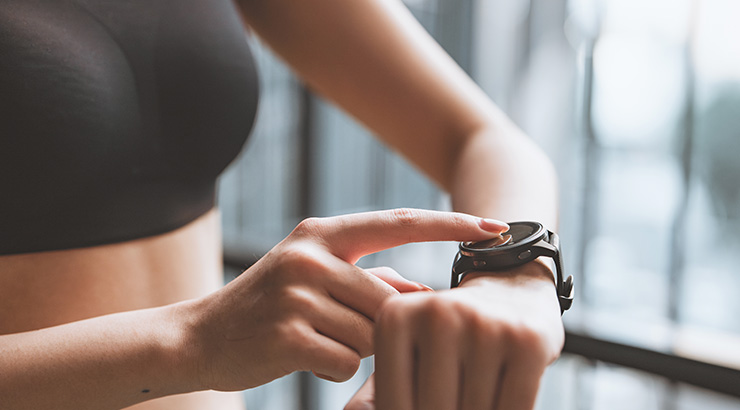 Kobieta dotyka palcem czarnego smartwatcha na nadgarstku. W tle okno z rozmytym widokiem. Ma na sobie czarny sportowy top.