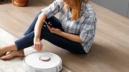 Kobieta siedzi na podłodze z telefonem, obok biały robot sprzątający na dywanie, w tle panele i meble.