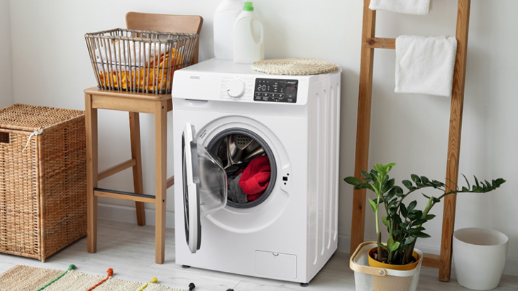 W łazience stoi biała pralka z otwartymi drzwiami, wewnątrz widać czerwone ubrania. Obok kosz na pranie i drewniana drabina z ręcznikami.