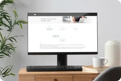 Na biurku stoi komputer z klawiaturą, obok kubek, notatnik i biały wazon. W tle widoczna roślina doniczkowa.