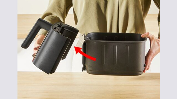 Osoba trzyma czarną obudowę frytkownicy beztłuszczowej nad pojemnikiem. Czerwona strzałka wskazuje miejsce mocowania.