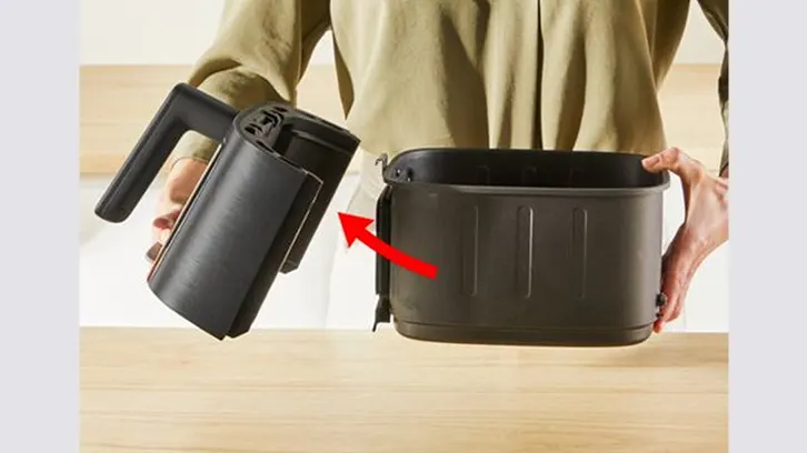 Osoba trzyma czarną obudowę frytkownicy beztłuszczowej nad pojemnikiem. Czerwona strzałka wskazuje miejsce mocowania.