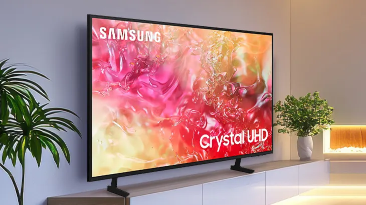 Telewizor Samsung z kolorowym ekranem stoi na szafce. Obok roślina doniczkowa i dekoracja. Z lewej strony widoczna druga roślina.