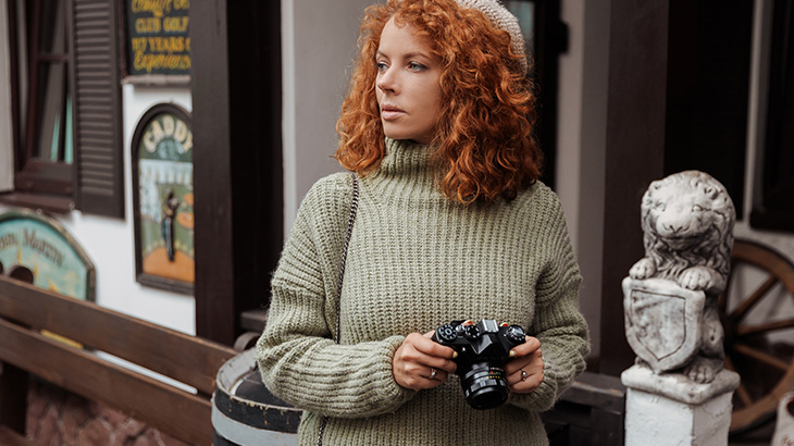 Kobieta w swetrze trzyma aparat, patrzy w bok. W tle budynek, rzeźba lwa i drewniane koło.