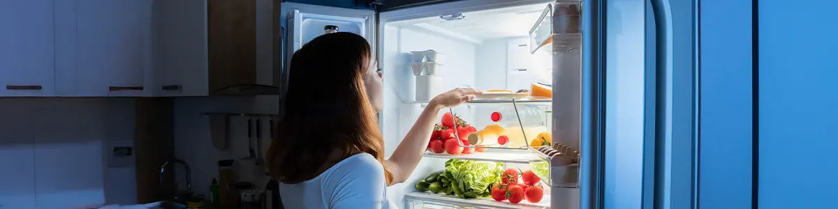 Kobieta w zielonej koszulce otwiera lodówkę pełną owoców i warzyw. W tle widać półki z kubkami i słoikami.