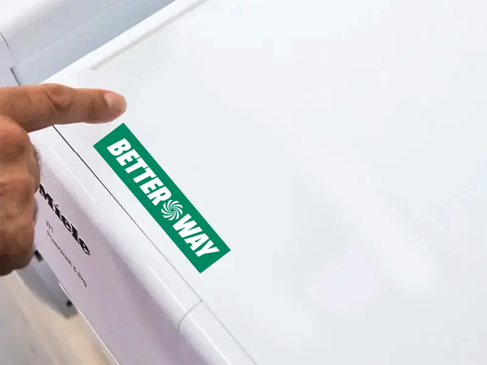 Palec wskazujący na białą pralkę Miele z zielonym logo "Better Way".