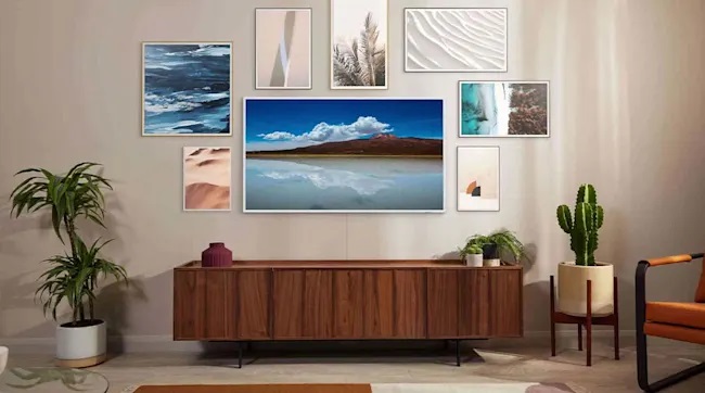 Pokój z roślinami i drewnianą szafką pod telewizorem. Na ścianie wisi telewizor i obrazy w białych ramkach.