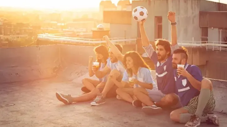 Grupa osób siedzi na dachu, świętując z piłką nożną i napojami, budynki w tle.