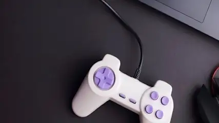 Na ciemnym tle leżą biały kontroler z fioletowymi przyciskami, czarna mysz z czerwonym kablem i fragment szarego laptopa.
