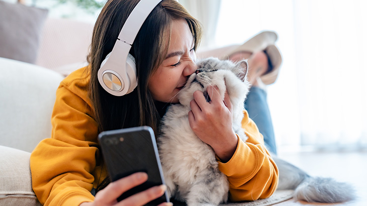 Kobieta w żółtej bluzie i słuchawkach przytula szarego kota, trzymając telefon. W tle widać białą sofę i okno.