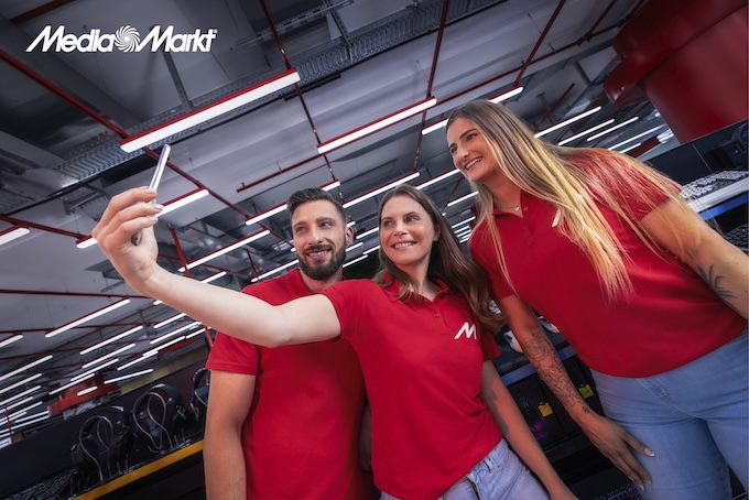 Trzy osoby w czerwonych koszulkach robią selfie w sklepie z oświetleniem i sprzętem elektronicznym. W tle widoczne logo Media Markt.