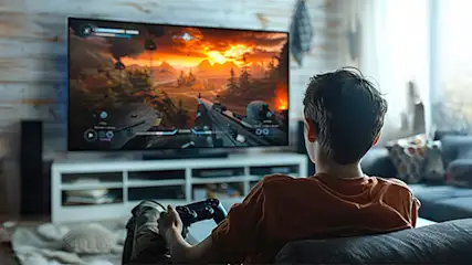 Chłopiec gra w grę wideo na telewizorze, siedząc na kanapie z kontrolerem w rękach. W tle widać okno i rośliny.