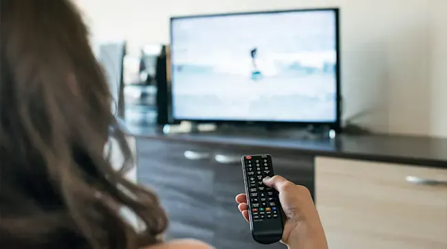 Osoba trzyma czarny pilot do telewizora. Telewizor stoi na szafce RTV w odcieniach brązu i beżu.