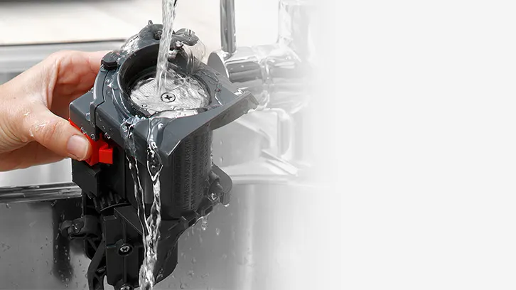 Osoba myje pod bieżącą wodą szary element ekspresu do kawy nad metalowym zlewem, trzymając go za czerwony uchwyt.