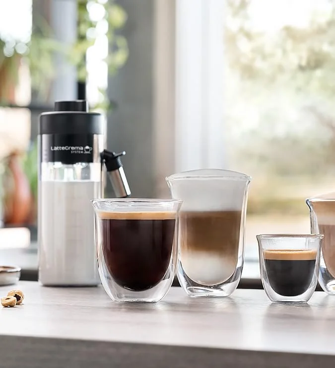 Na blacie stoją szklanki z kawą i mlekiem, obok pojemnik LatteCrema, w tle okno i rośliny.